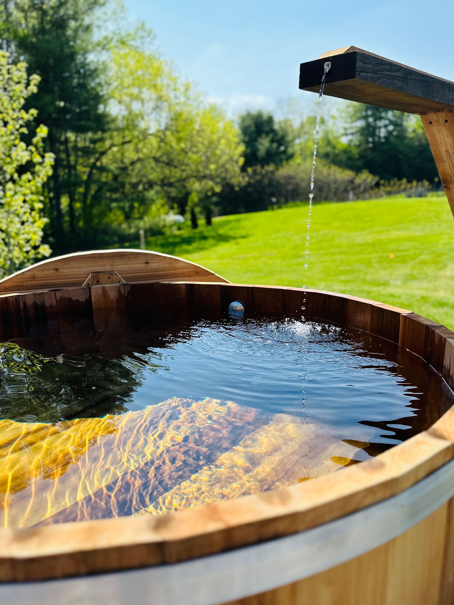 Wooden hot tub with water feature, set outdoors on a sunny day.