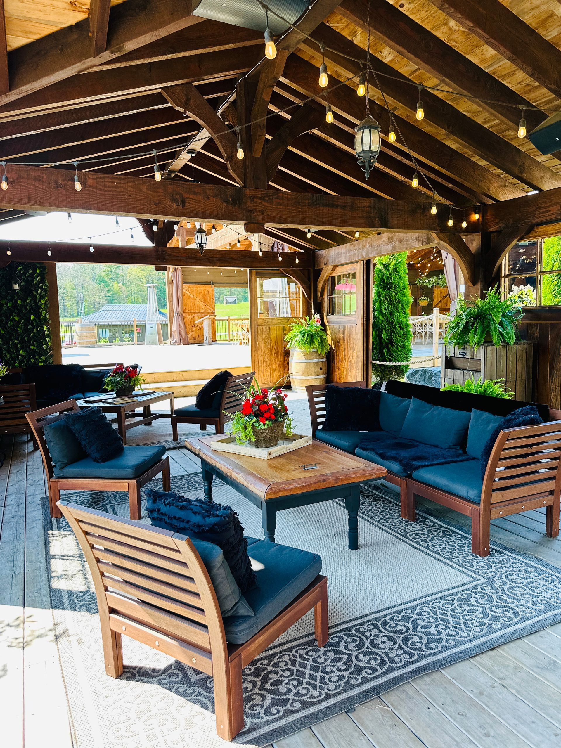 Outdoor seating area under a wooden roof. Blue cushions on wooden furniture, with a rug and string lights.