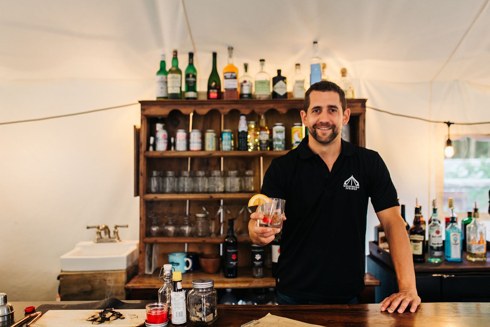 Bartender smiles, holding a cocktail at outdoor bar. Shelves with bottles and glassware.