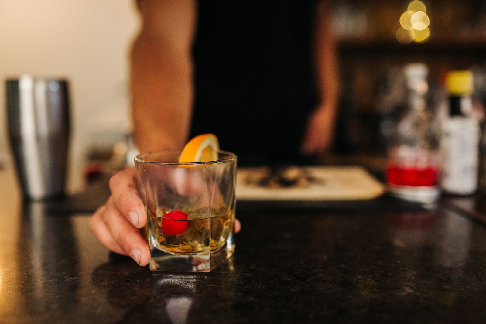 Bartender offers an Old Fashioned cocktail with orange slice and cherry garnish on a dark bar.