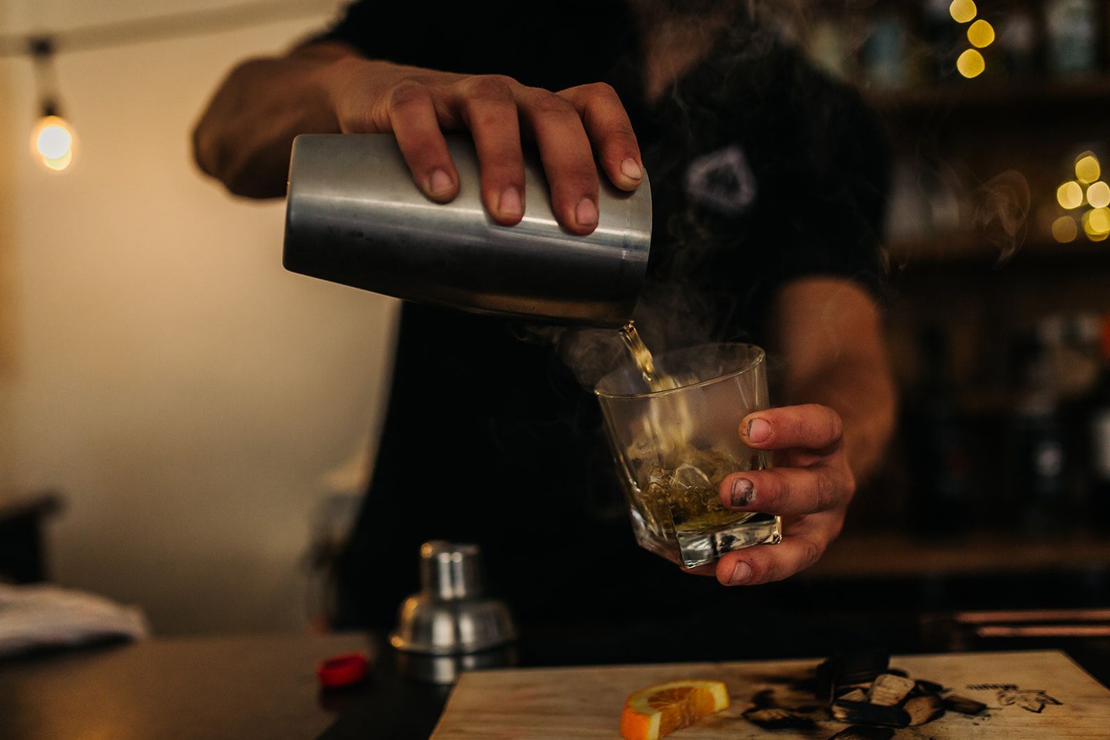 Bartender pours drink from shaker into a rocks glass, steam rising. Lit bar setting, close up.