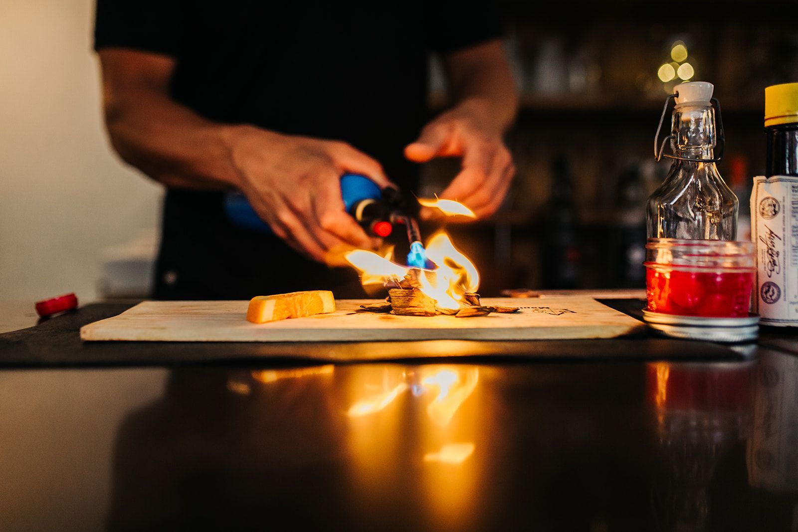 Bartender torches a food item on a wooden board; flames and bottled ingredients on a dark bar.