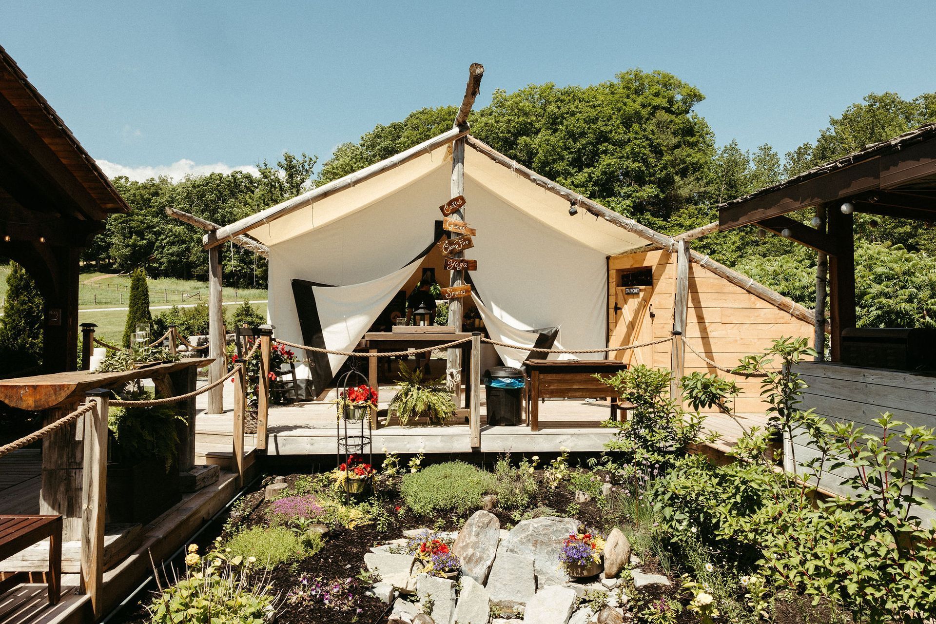 Glamping tent on wooden deck, flowers in foreground, blue sky, trees in background.