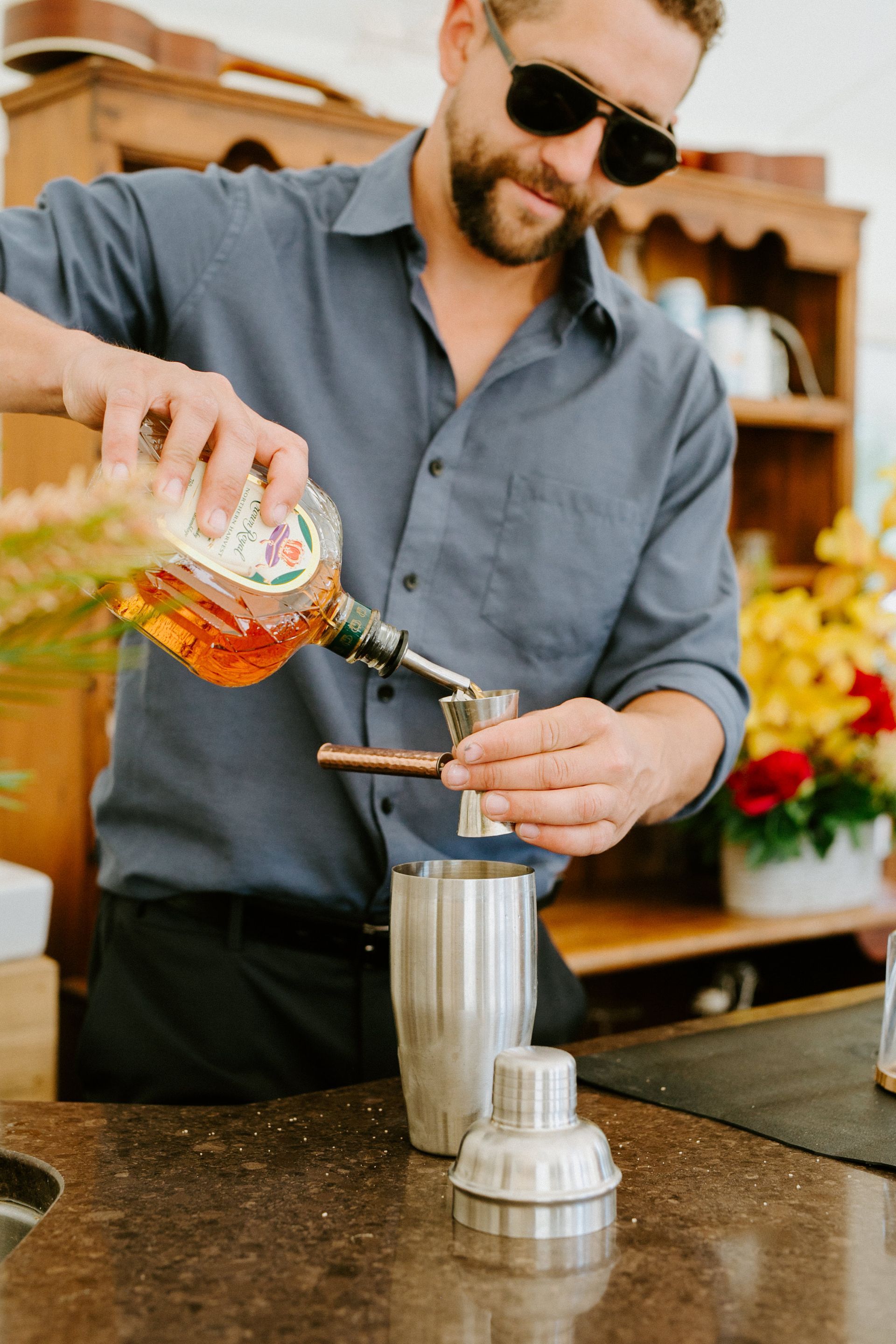 Bartender pouring liquid from a bottle into a jigger over a cocktail shaker, outdoors.