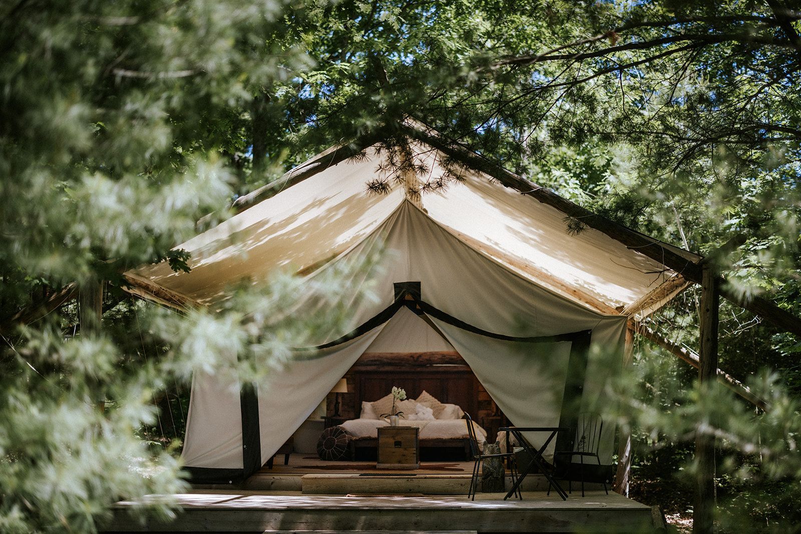 Glamping tent nestled in trees; cream canvas, wooden frame, bed visible inside.