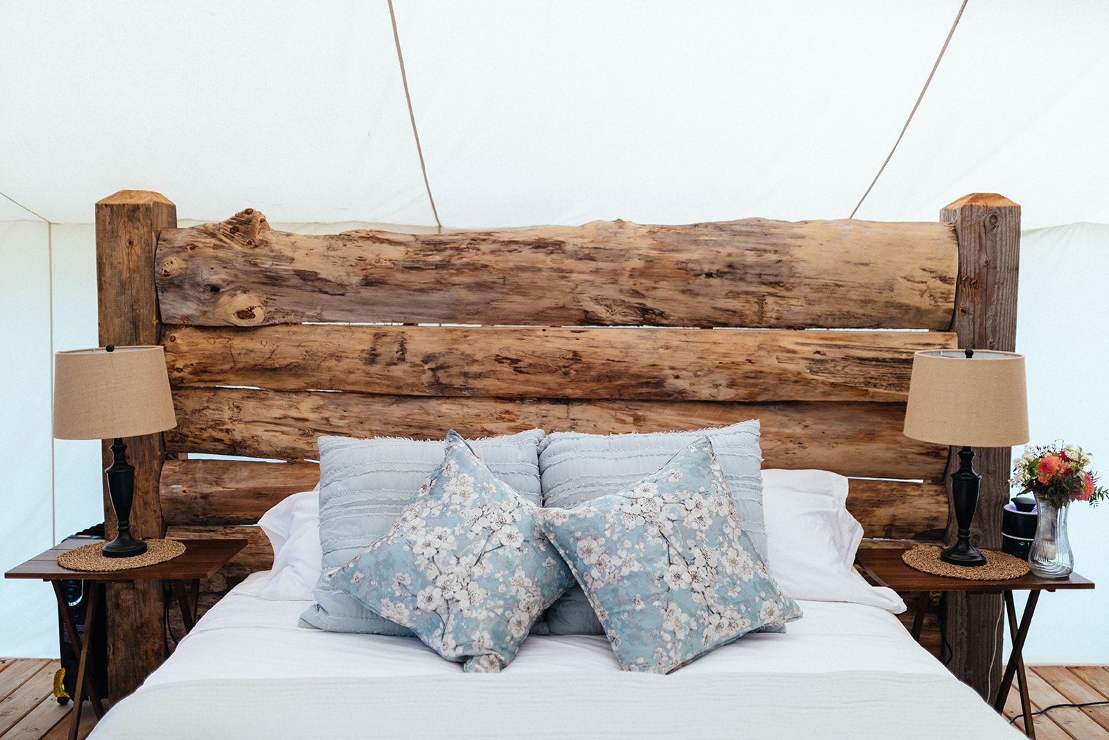 Bed with rustic log headboard, bedside tables with lamps, light blue pillows, flower vase, inside a tent.