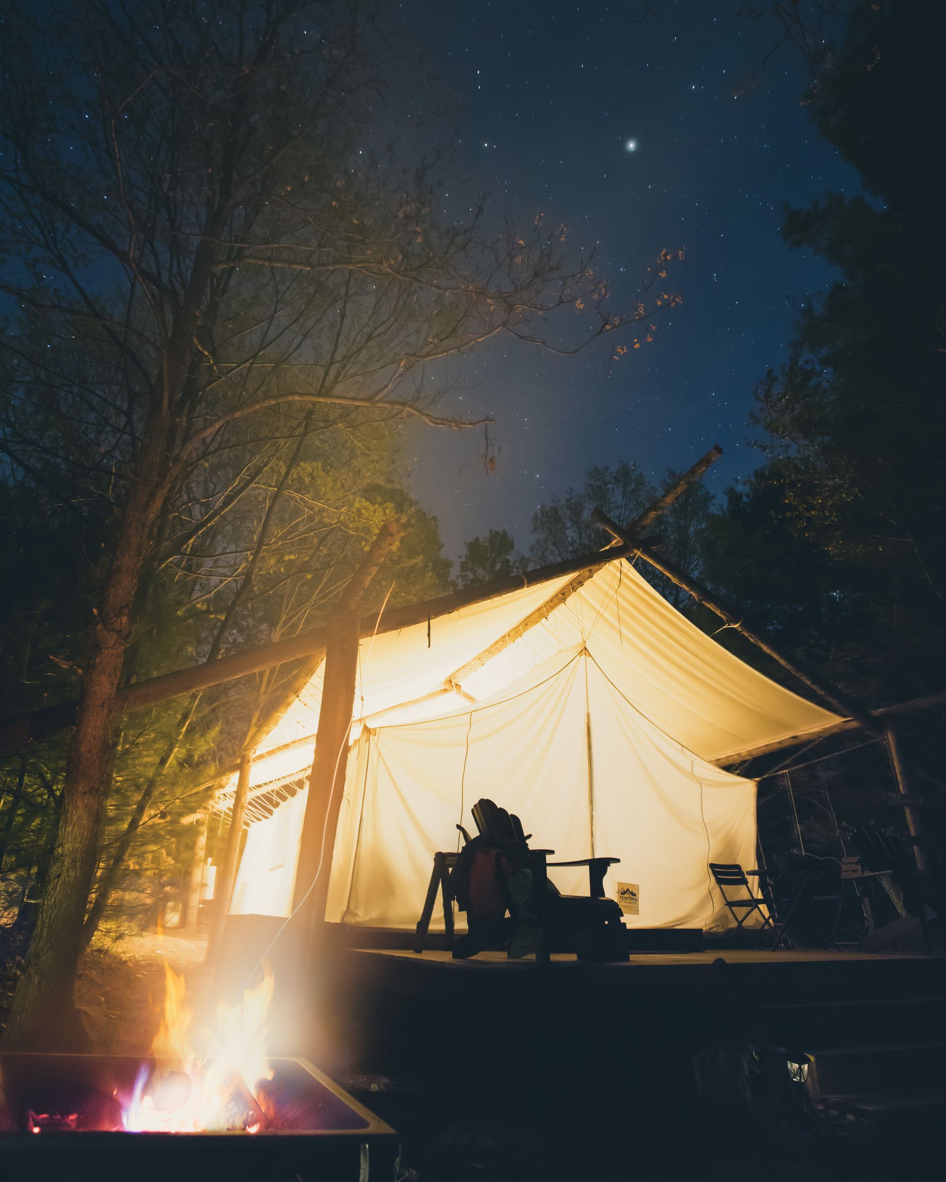 Lit campsite at night. Person sitting in a chair, reading under a glowing tent. Fire in the foreground, stars overhead.