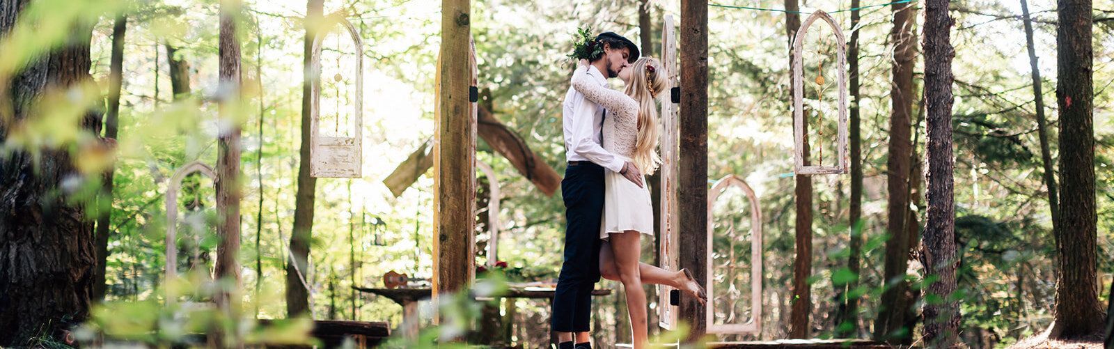Couple embracing, kissing in a forest, surrounded by trees. Man in a white shirt and black pants, woman in a white dress.