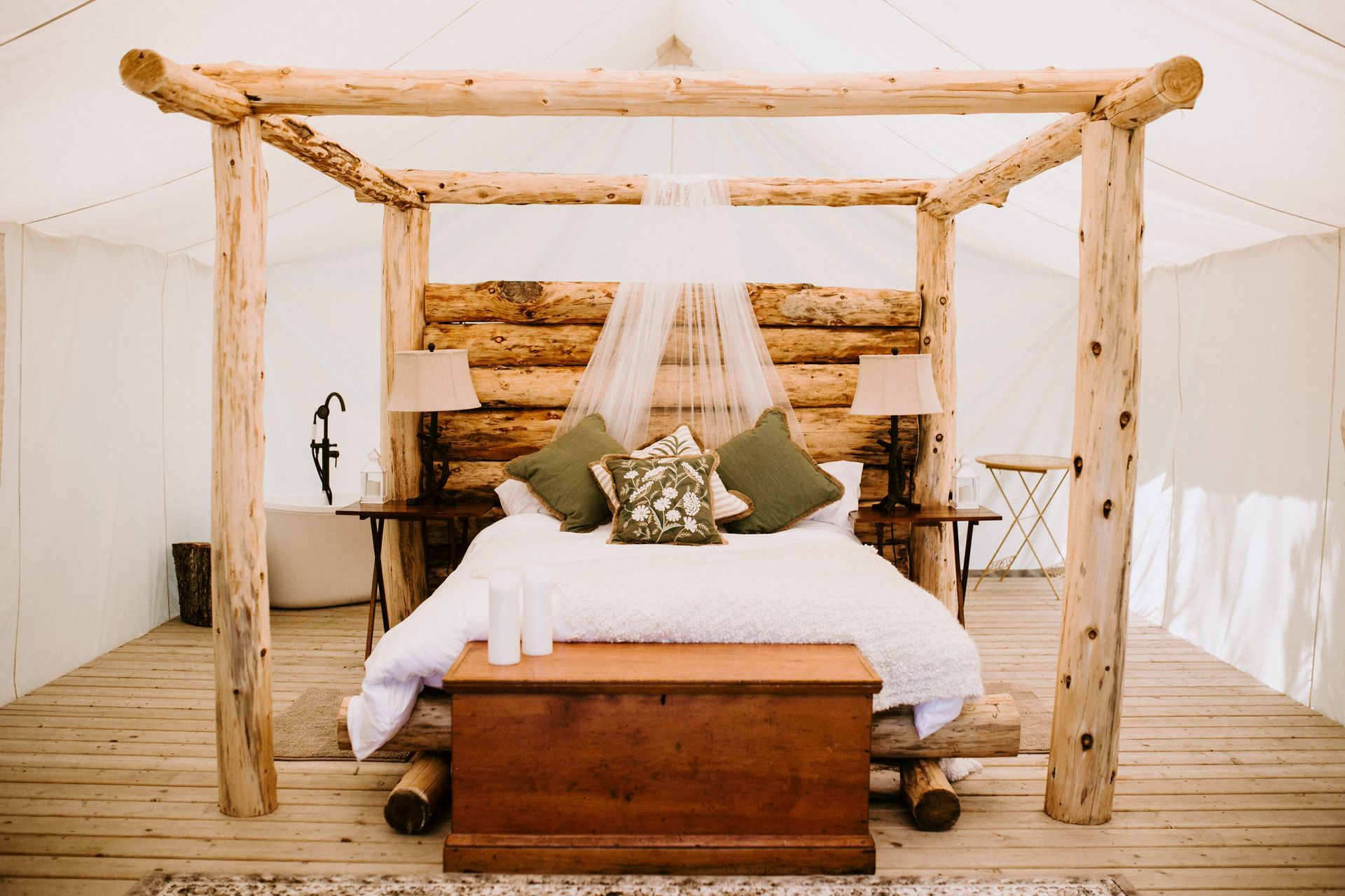 Rustic bedroom with log-framed bed, wooden headboard, canopy, and trunk at foot. Neutral tones.