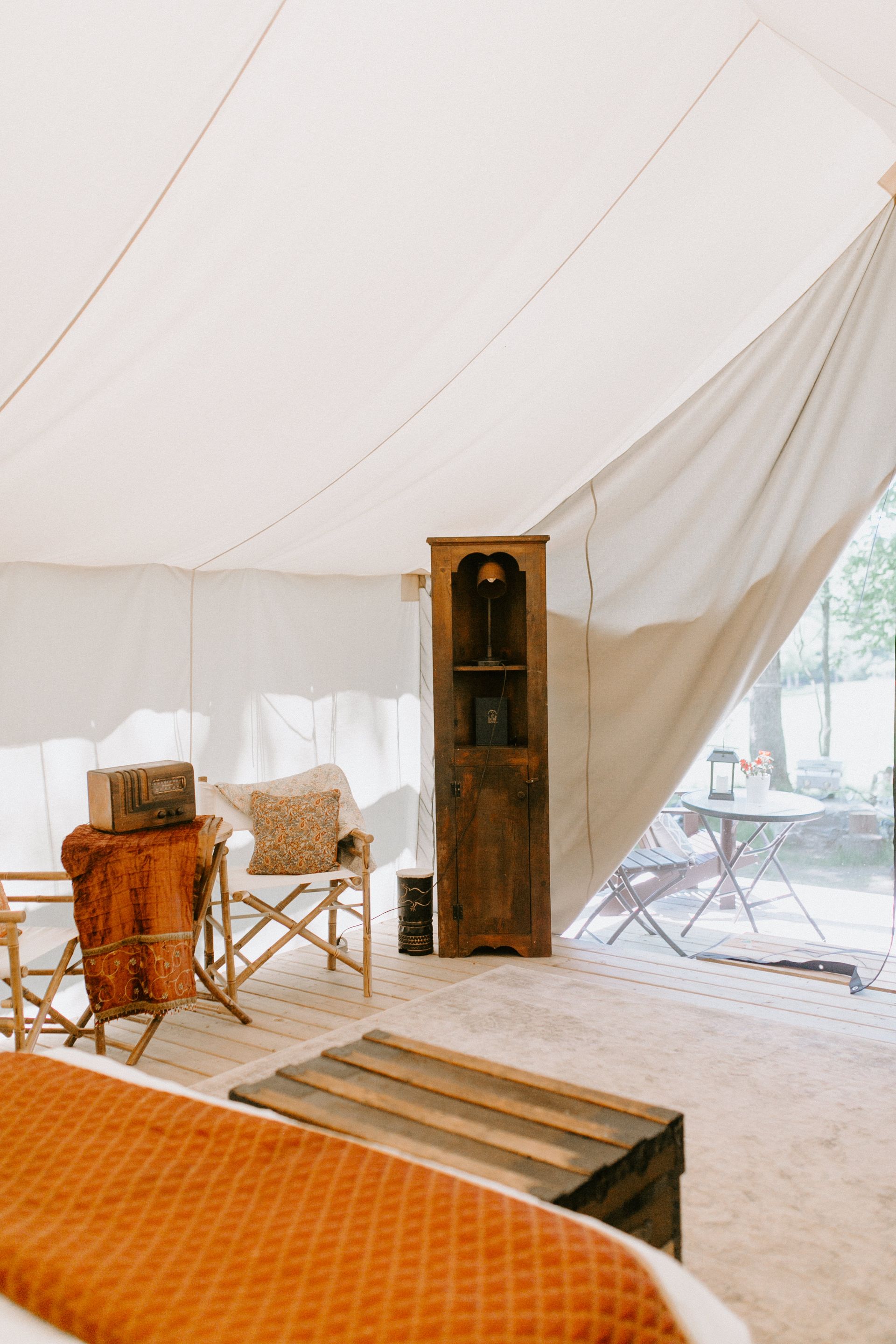 Interior of a canvas tent with furniture: bed, chair, cabinet, rug. Sunlight streams in.