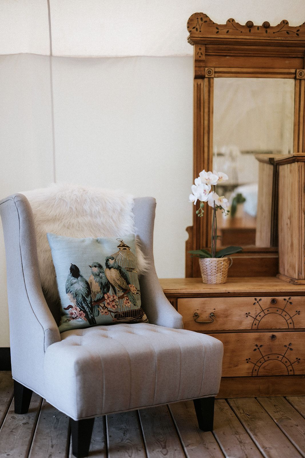 A gray upholstered armchair with a pillow and a sheepskin throw is next to an antique wooden dresser with a mirror.