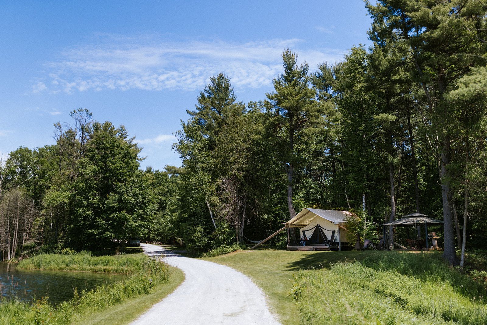 Gravel road curves to a small, yellow building nestled in a grove of trees under a blue sky.