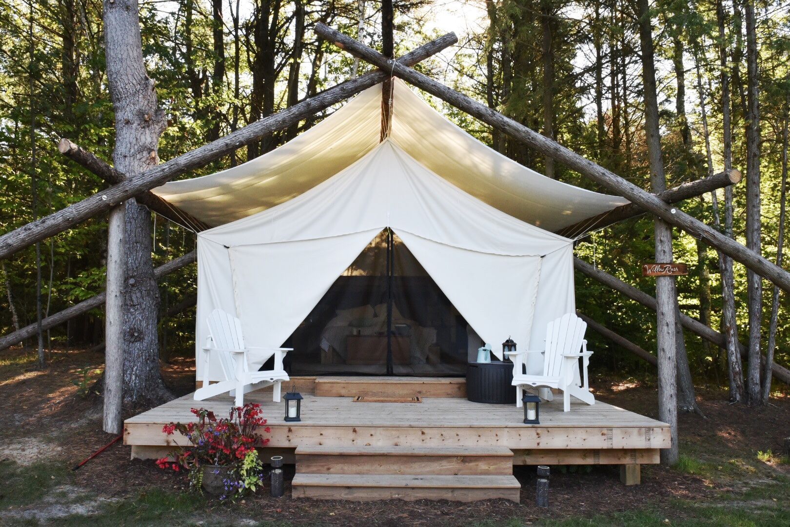 White canvas glamping tent on a wooden platform in a forest, with chairs and lanterns.