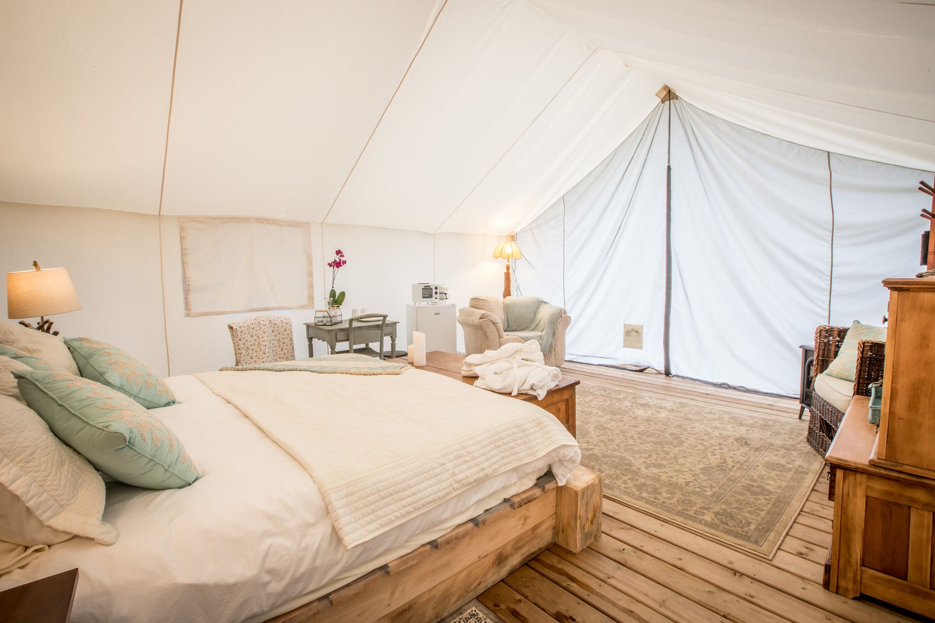 Interior of a canvas tent with a bed, seating area, wooden furniture, and neutral decor.