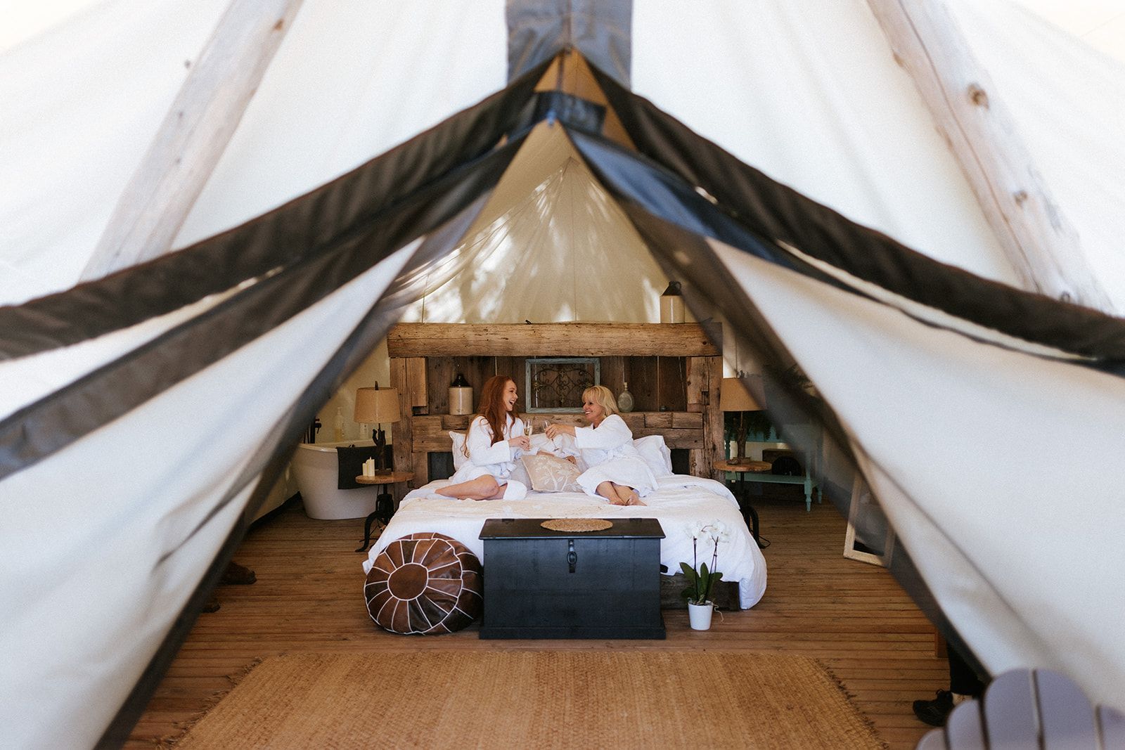 Two people in white robes relax in a glamping tent with a bed and wooden headboard.