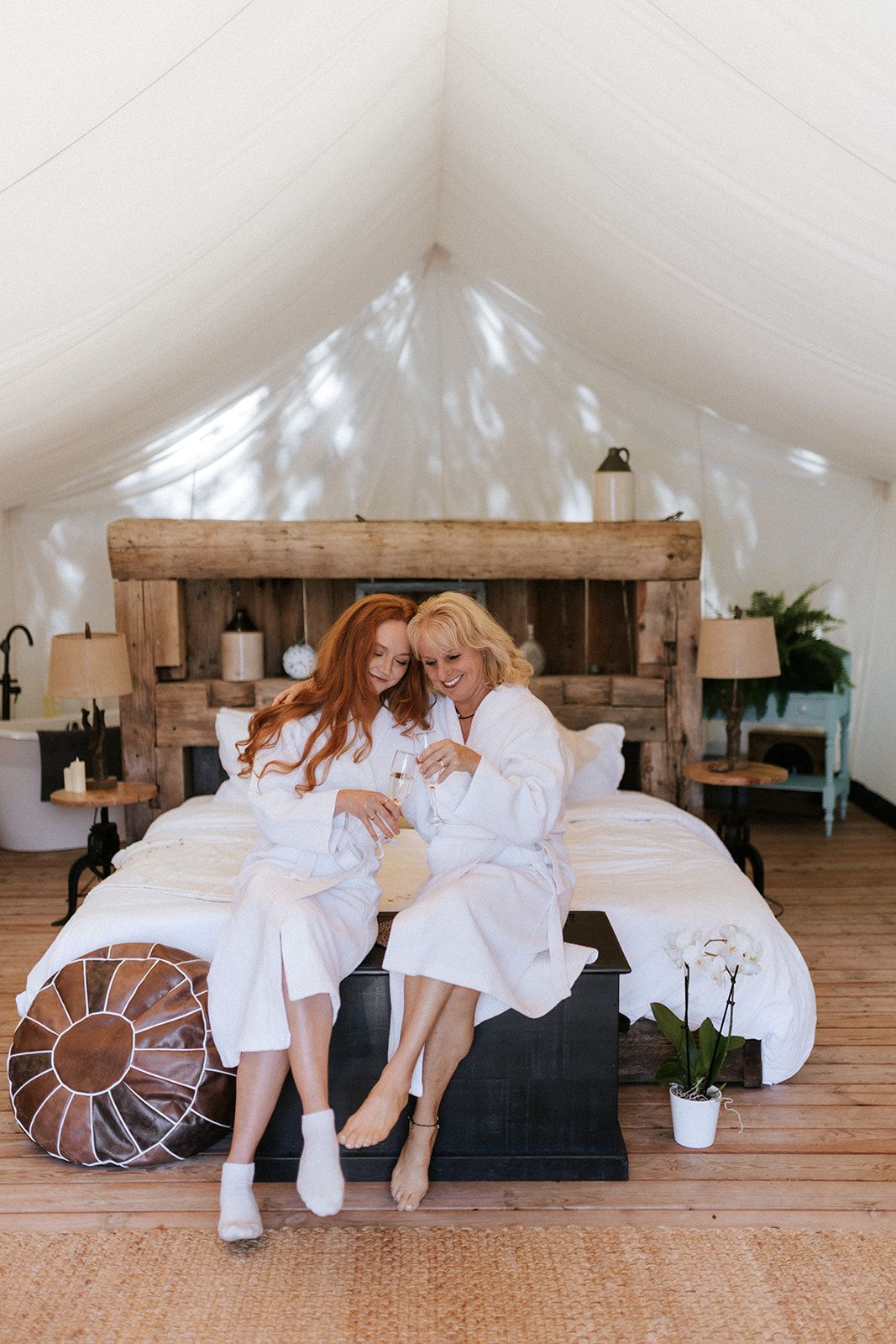 Two people in white robes sit on a bed in a tent-like room; one with red hair, the other blonde.