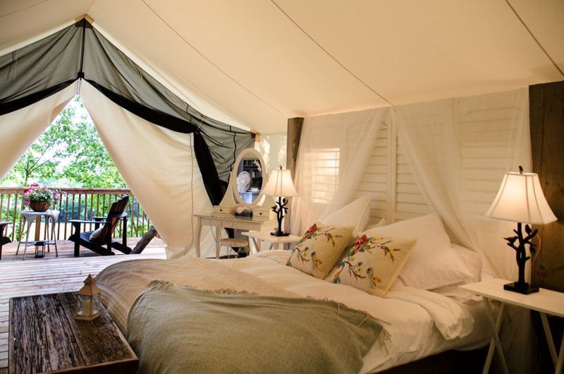 Inside of a luxury tent: white walls, large bed, vanity table, and open to a deck with chairs.