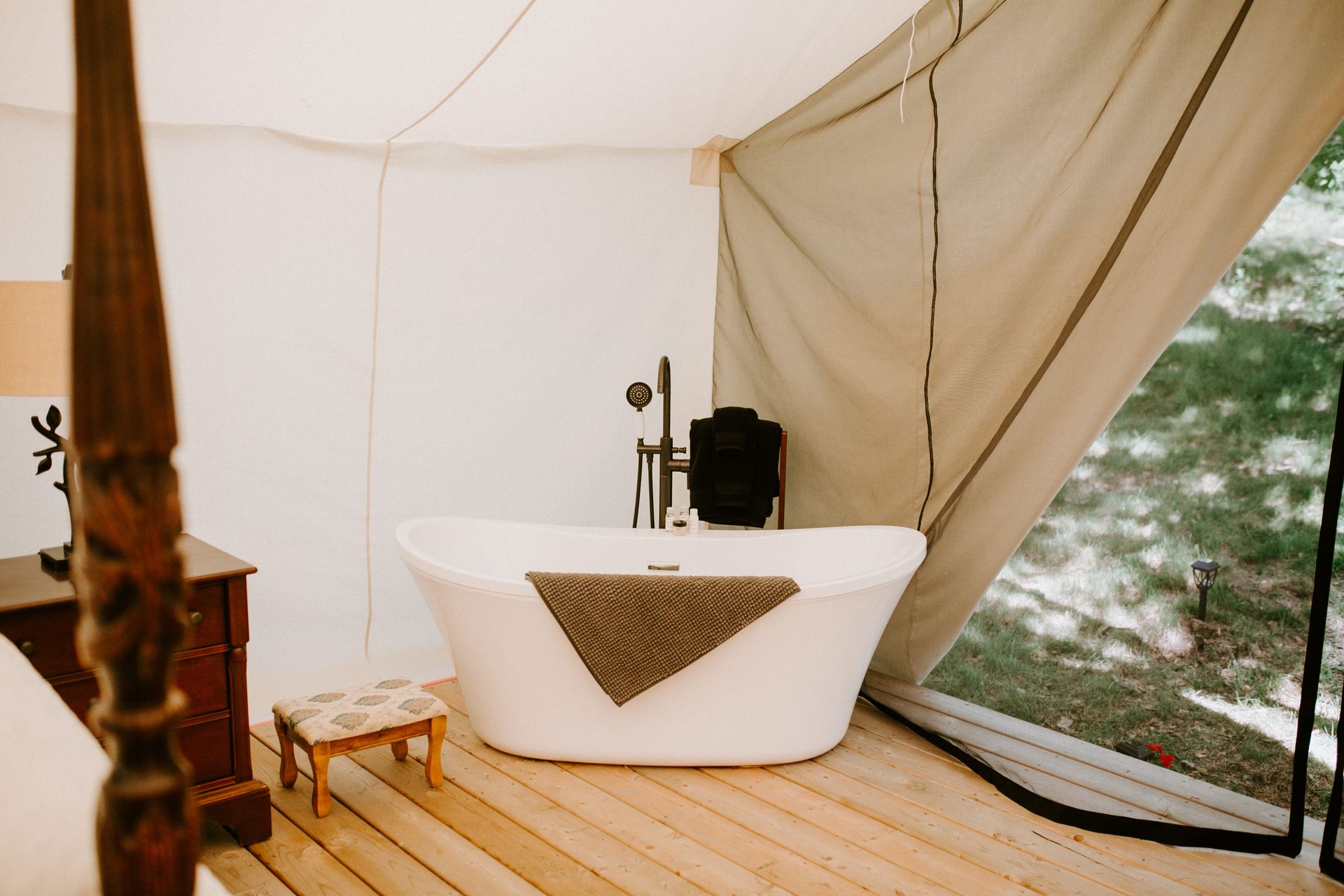 Luxury glamping tent interior with freestanding white bathtub, wooden floor, and open flap to the outside.