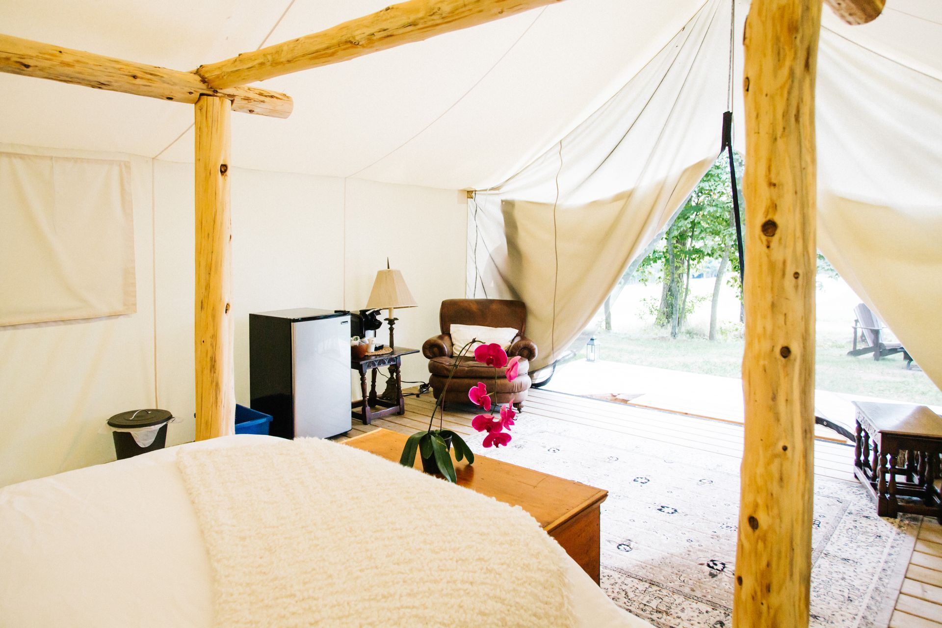 Interior of a glamping tent with a bed, small fridge, armchair, and table. White canvas walls, wooden beams.