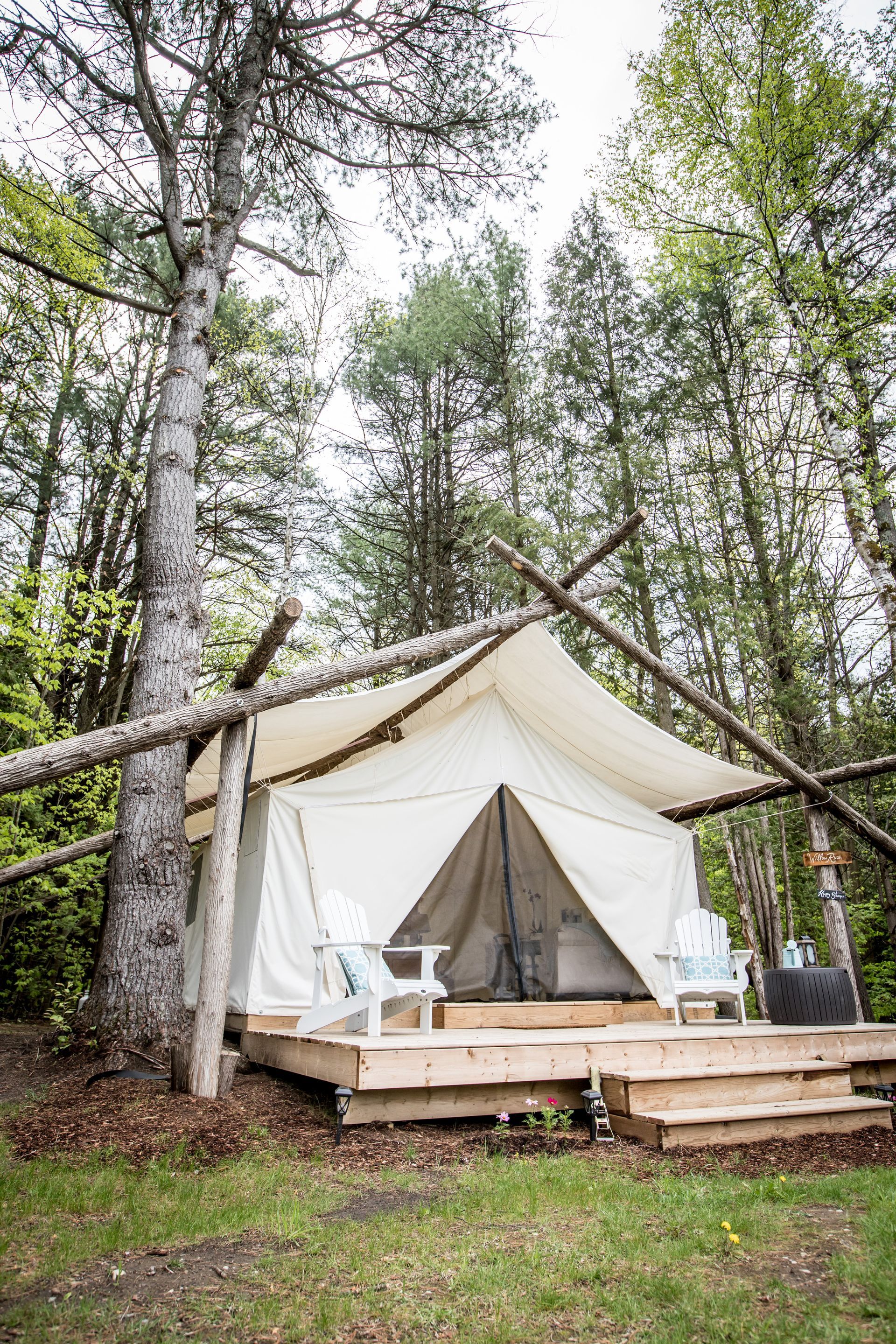 Canvas tent on wooden deck, supported by logs, in a forest. Two white chairs sit outside.