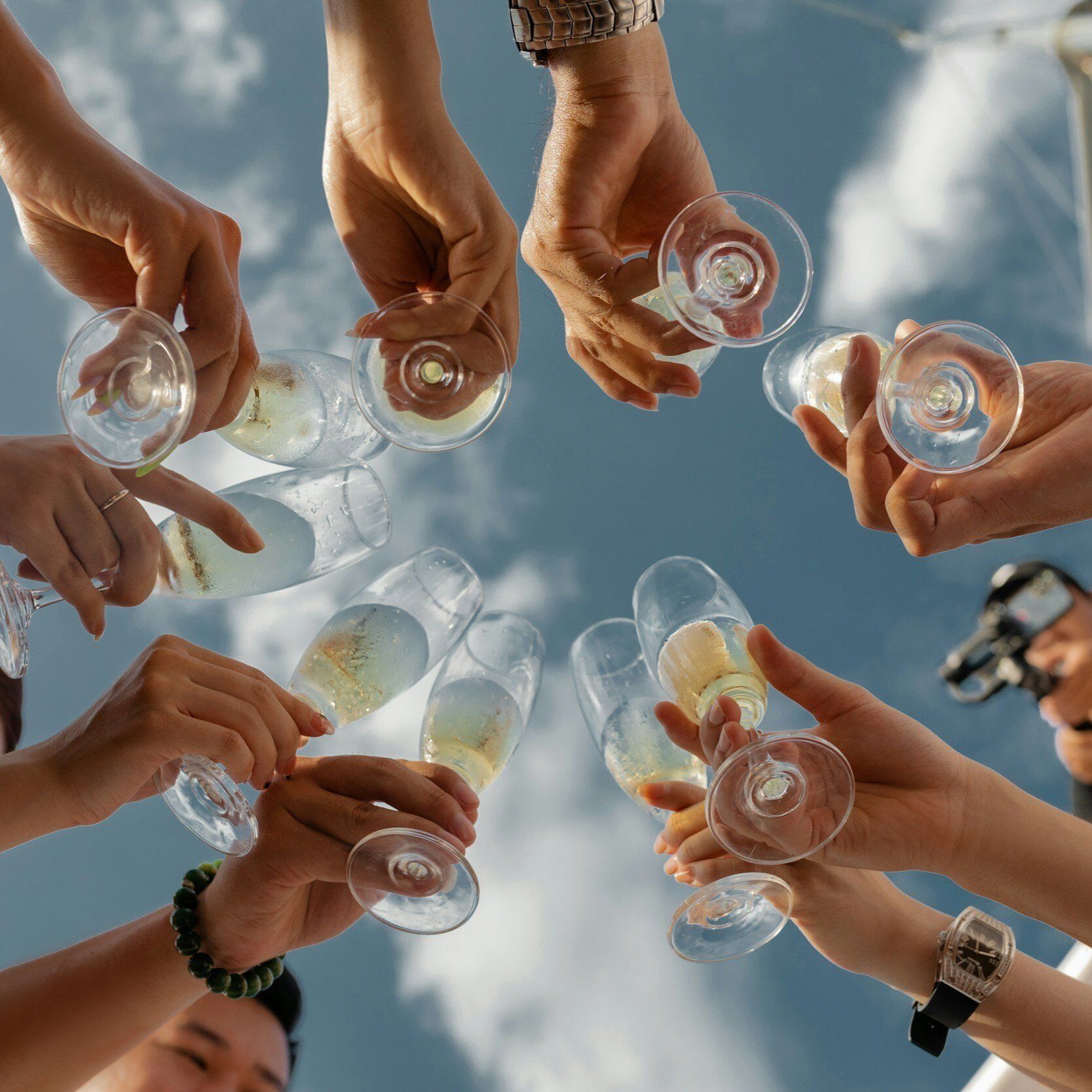 Hands clinking champagne glasses in a toast against a blue sky with clouds.