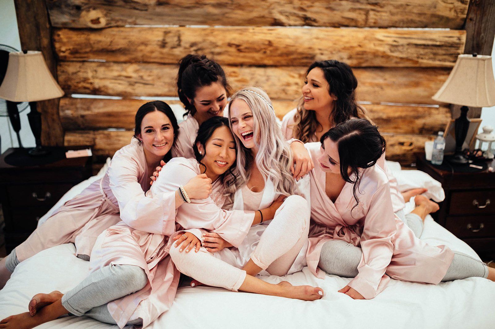 Bridesmaids and bride laugh together, wearing robes, on a bed with a wooden headboard.