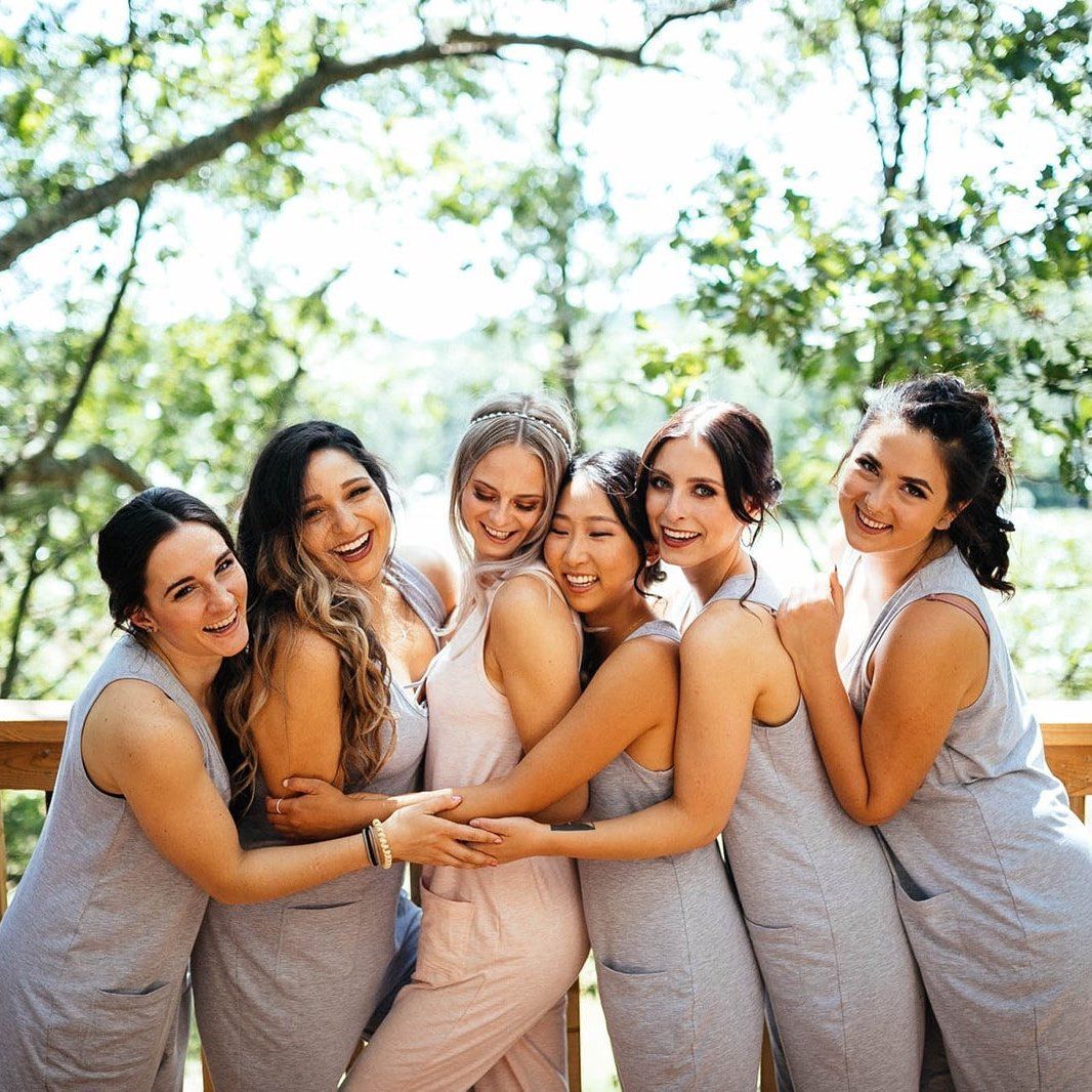 Bridesmaids in gray dresses hugging the bride in a blush jumpsuit on a wooden deck.