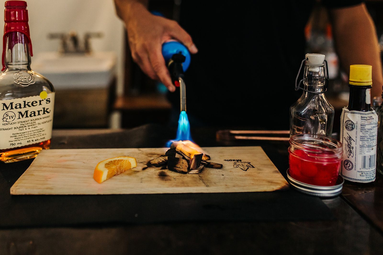 Bartender torches wood with orange slice, cocktail ingredients in background.