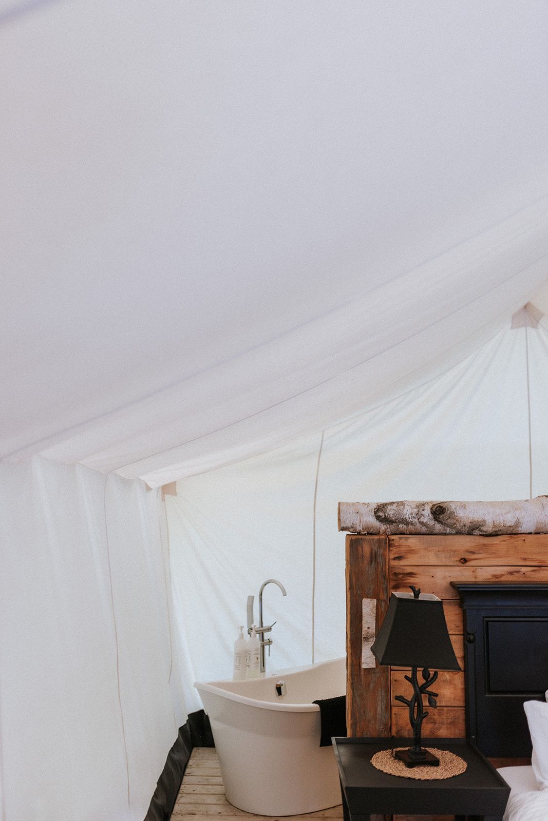 Interior of a glamping tent with a bathtub and a bed, featuring white canvas walls and a rustic wood headboard.