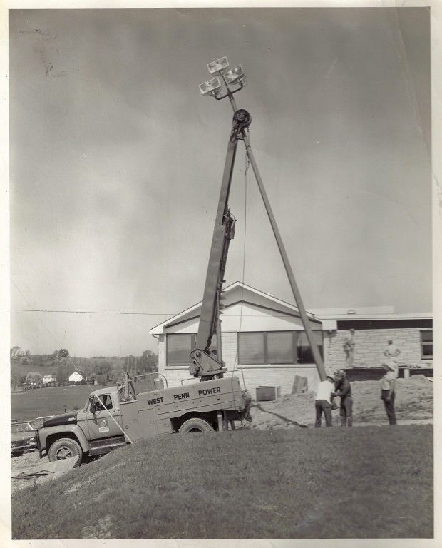 A black and white photo of a crane lifting a pole in front of a building.