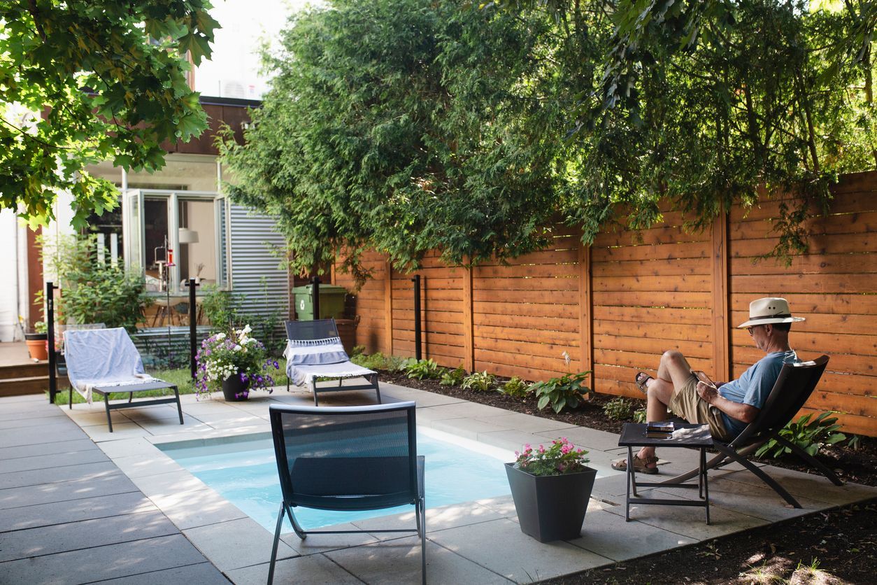 A man is sitting in a chair in a fenced backyard next to a pool.