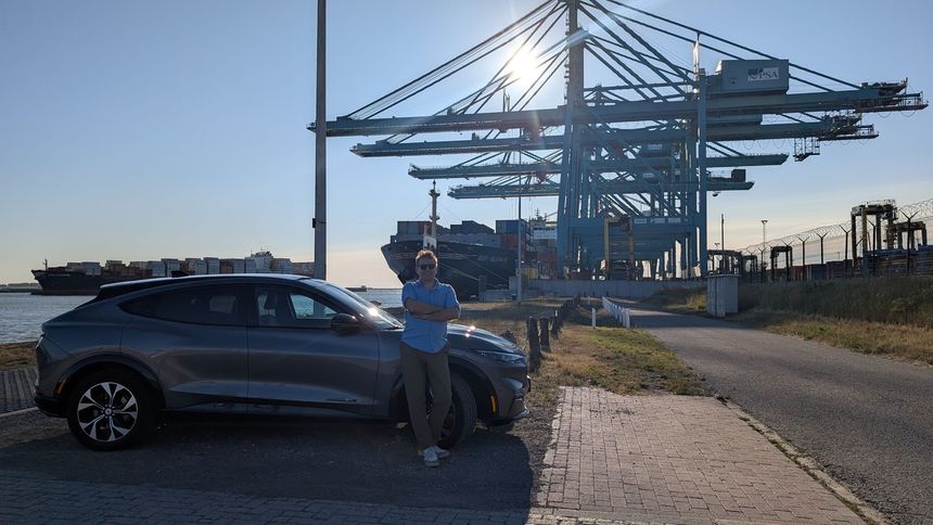 Man stands by a gray electric SUV at a port, arms crossed, harbor cranes and ship in background.