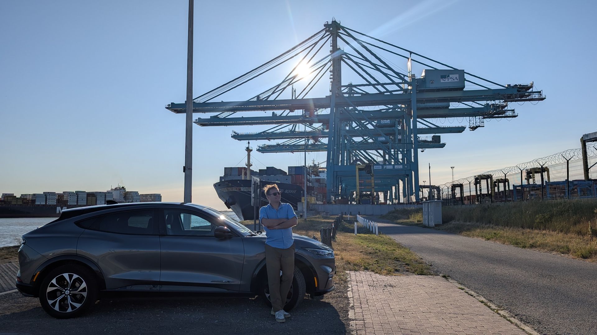 Een man staat bij een grijze elektrische SUV in een haven met kranen en een containerschip. Zonnig, blauwe lucht.