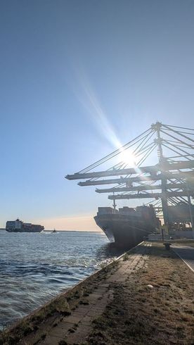 Sun shining over a container ship at a port; gantry cranes and another ship in the distance.