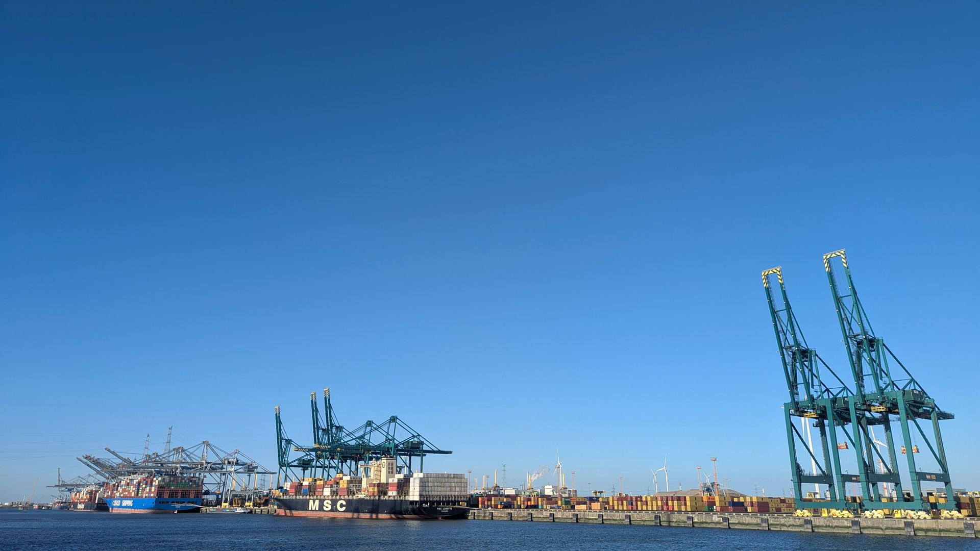 Blauer Himmel über einem geschäftigen Hafen mit Frachtkränen, Schiffen und Schiffscontainern.