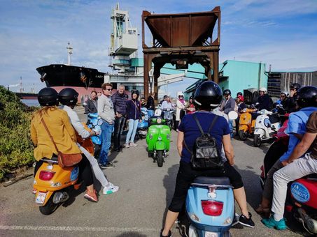 A group of people on colorful scooters, gathered near a harbor with a large ship and building in the background.
