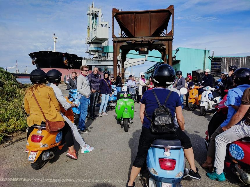 Man taking selfie with group of people on scooters in front of cargo ship at dock.