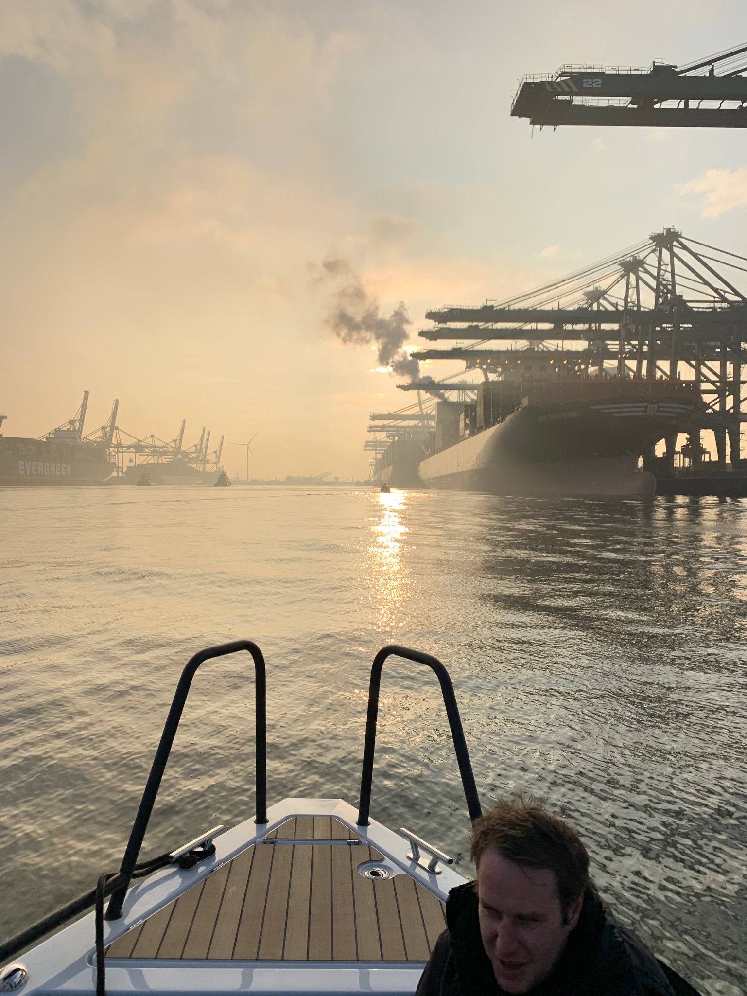 Bateau sur l'eau, face à un port avec des grues, coucher de soleil et un homme au premier plan.