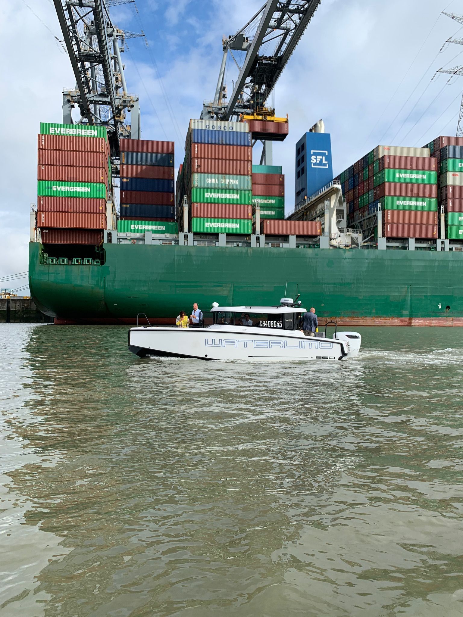 Un petit bateau blanc passe devant un grand cargo vert sous une grue à portique dans un port.