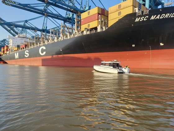 Small white boat on water in front of a large black and red cargo ship, MSC Madrid.