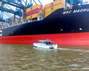 Small white boat on water in front of a large black and red cargo ship, MSC Madrid.
