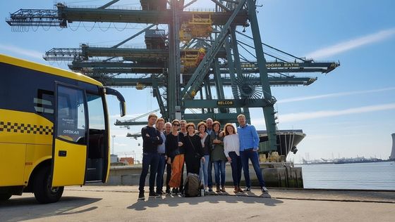A group stands by a yellow bus at a port with cranes and water.
