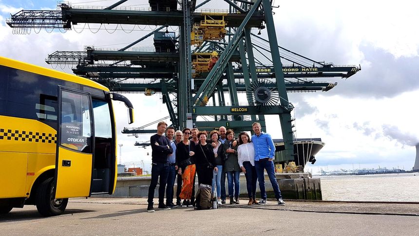 A group stands by a yellow bus at a port with cranes and water.