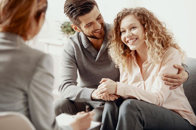 Couple smiling, holding hands, and embracing on a couch while meeting with a counselor.