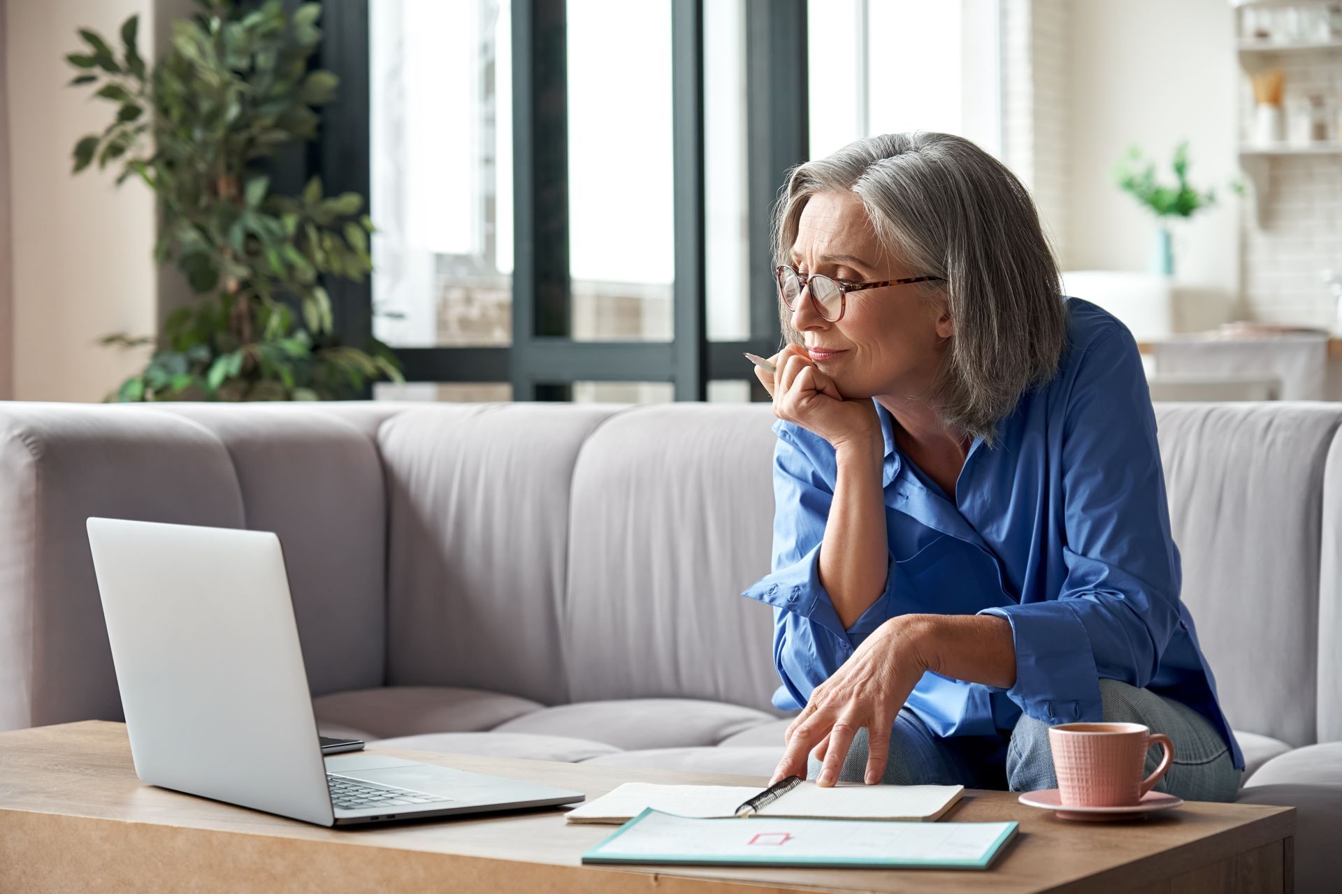 Senior woman with glasses, using laptop, resting chin on hand, looking at notes, living room setting.