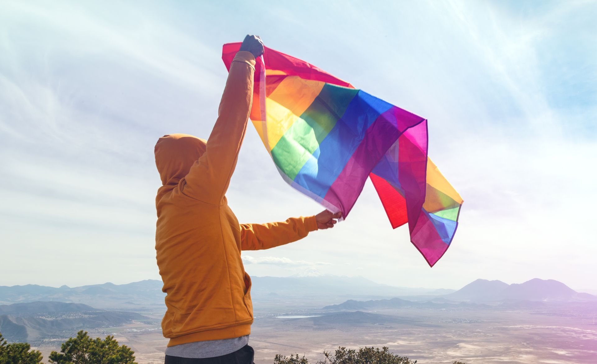 Person in orange hoodie holding rainbow flag aloft on hilltop overlooking mountains.