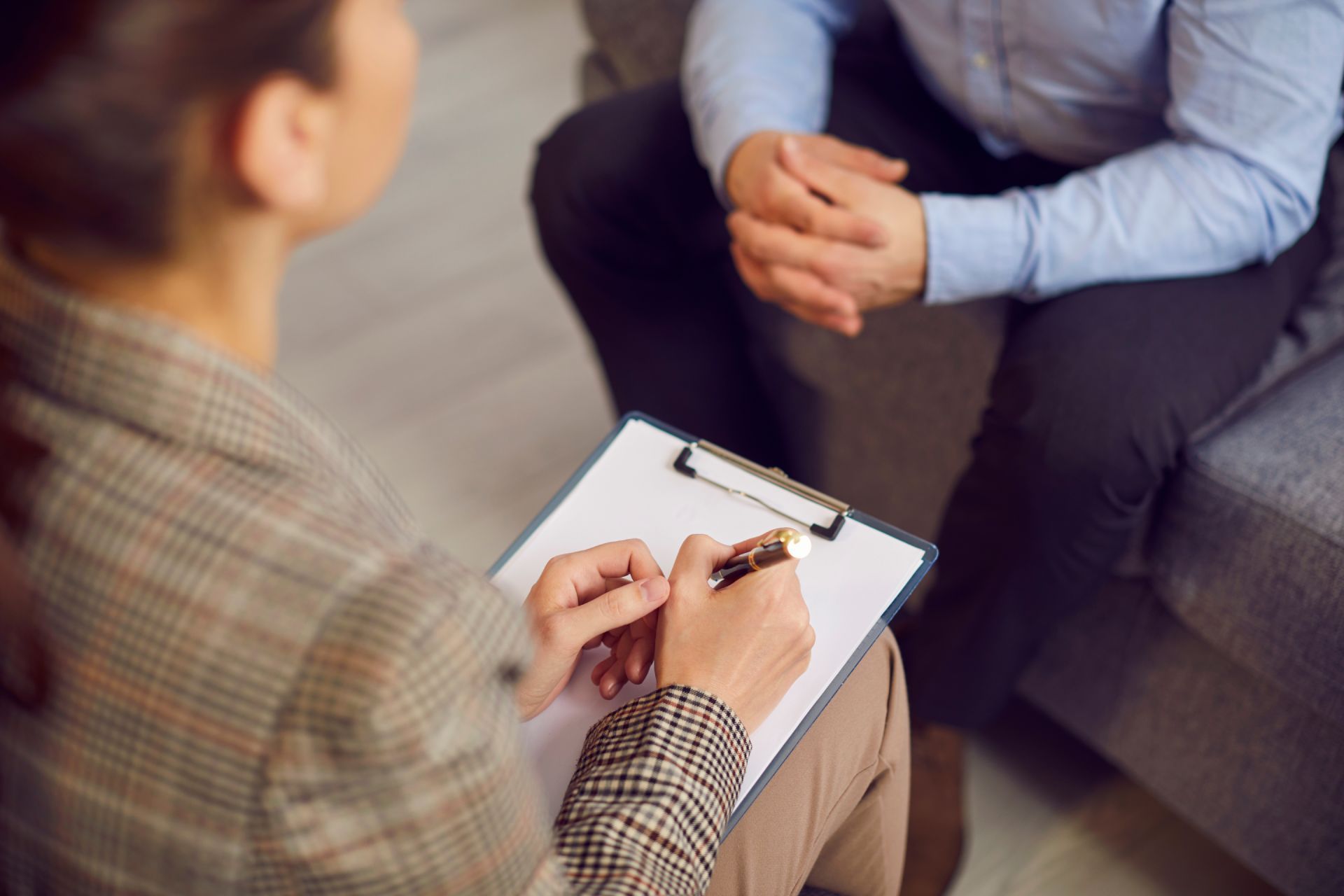 Therapist in plaid jacket taking notes while a man in a blue shirt sits on a couch.