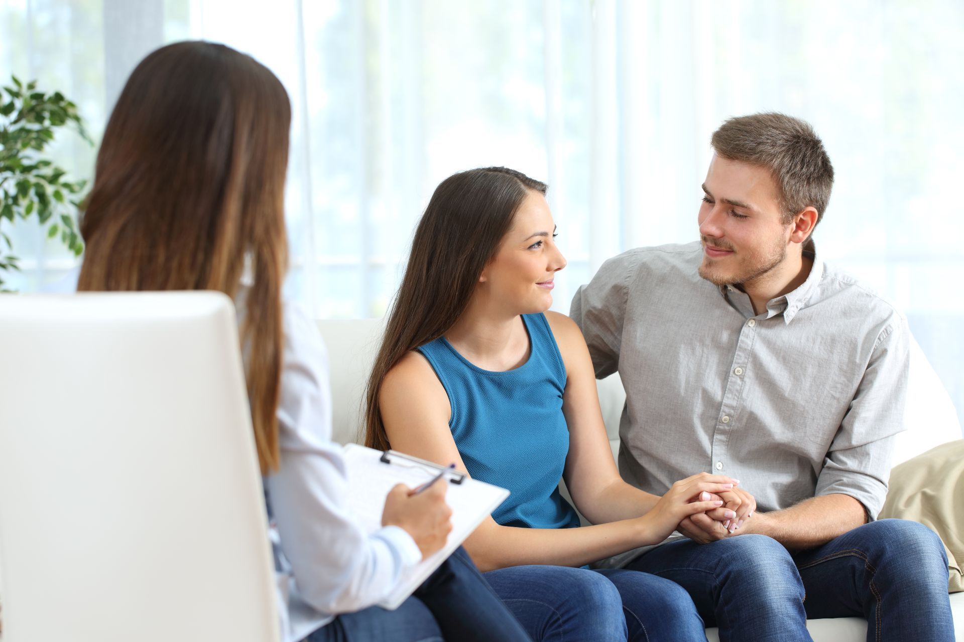 Couple holding hands and looking at each other during a counseling session; therapist taking notes.