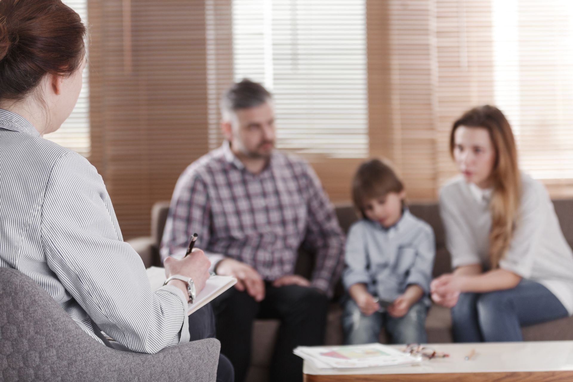 Therapist taking notes during a family therapy session. A sad child and parents sit on a couch.