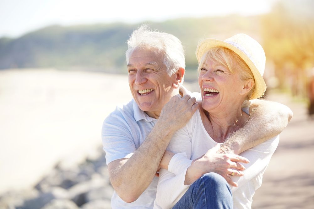 Elderly couple laughing, hugging outdoors by a sunny beach, man's arm around woman wearing a straw hat.